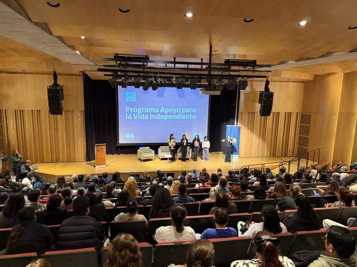 Auditorio con gran asistencia de público durante el lanzamiento de la capacitación de Asistentes Personales para la Vida Independiente, con expositoras en el escenario y pantalla del programa al fondo.